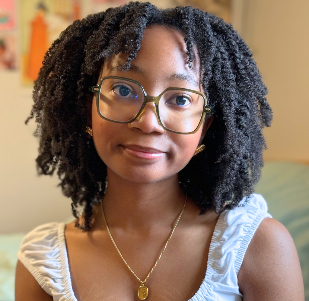 Close-up of a smiling woman with glasses, curly black hair, wearing a light blue ruffled top and a gold pendant necklace.