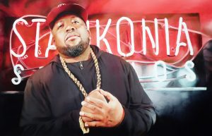 Man in a black outfit with a thick gold chain, standing in front of a red neon sign, facing the camera.