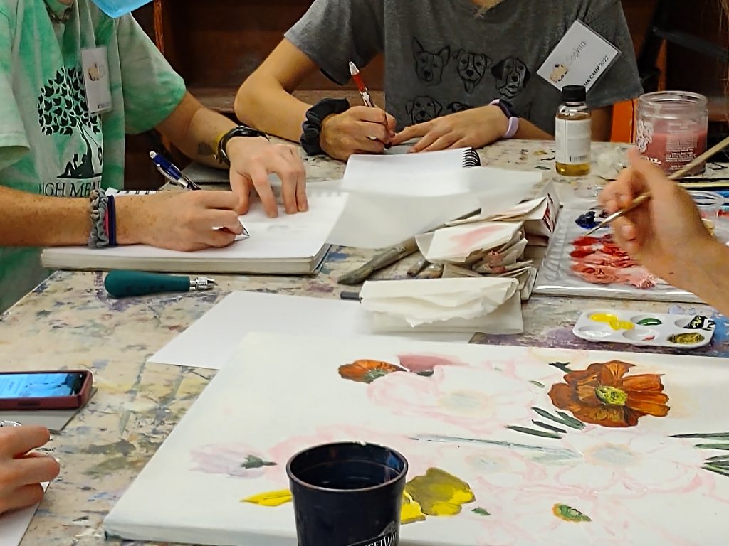 People sketching and painting at a crowded table with brushes, paper, and paint palettes.