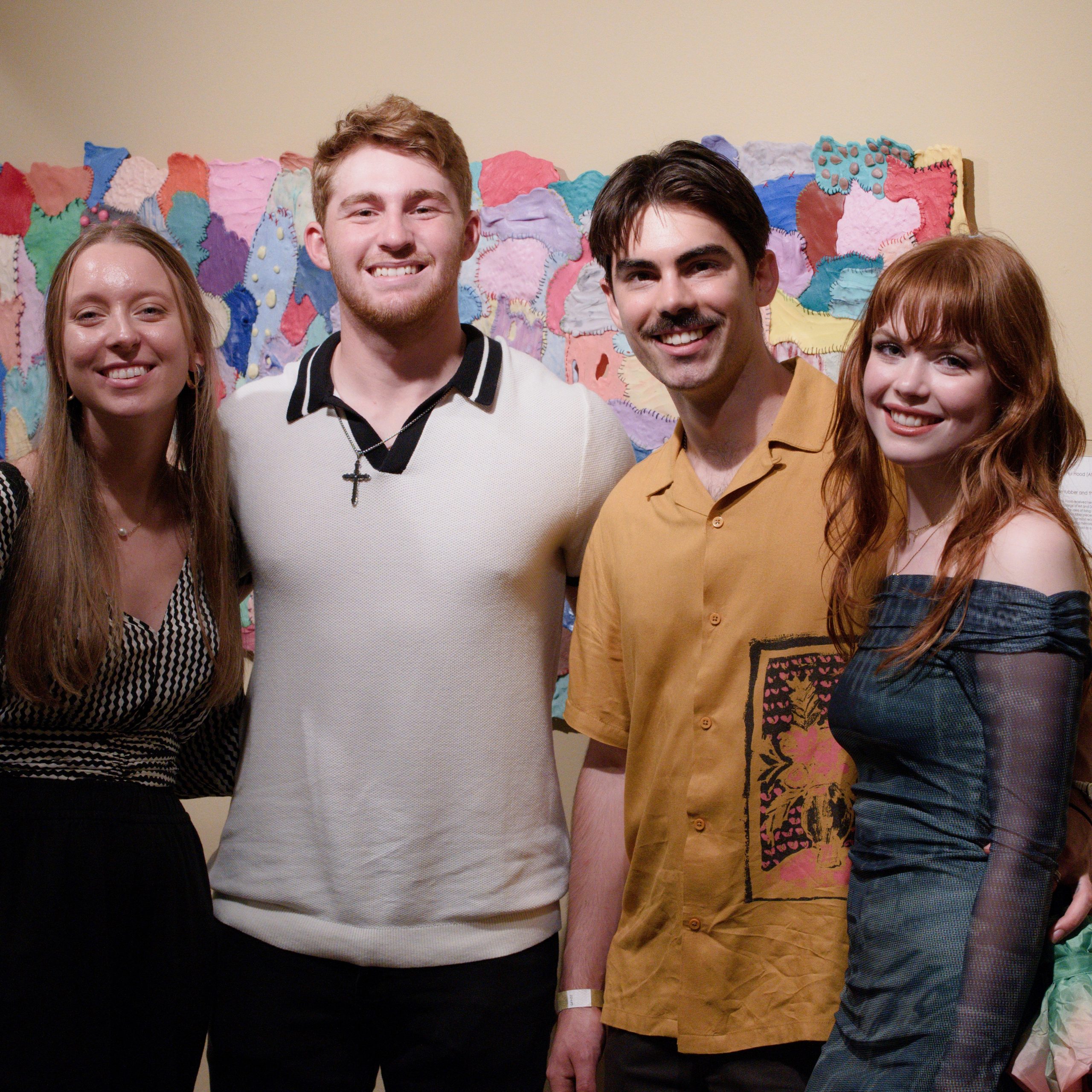 Four people posing indoors against a bright multicolored patchwork wall.
