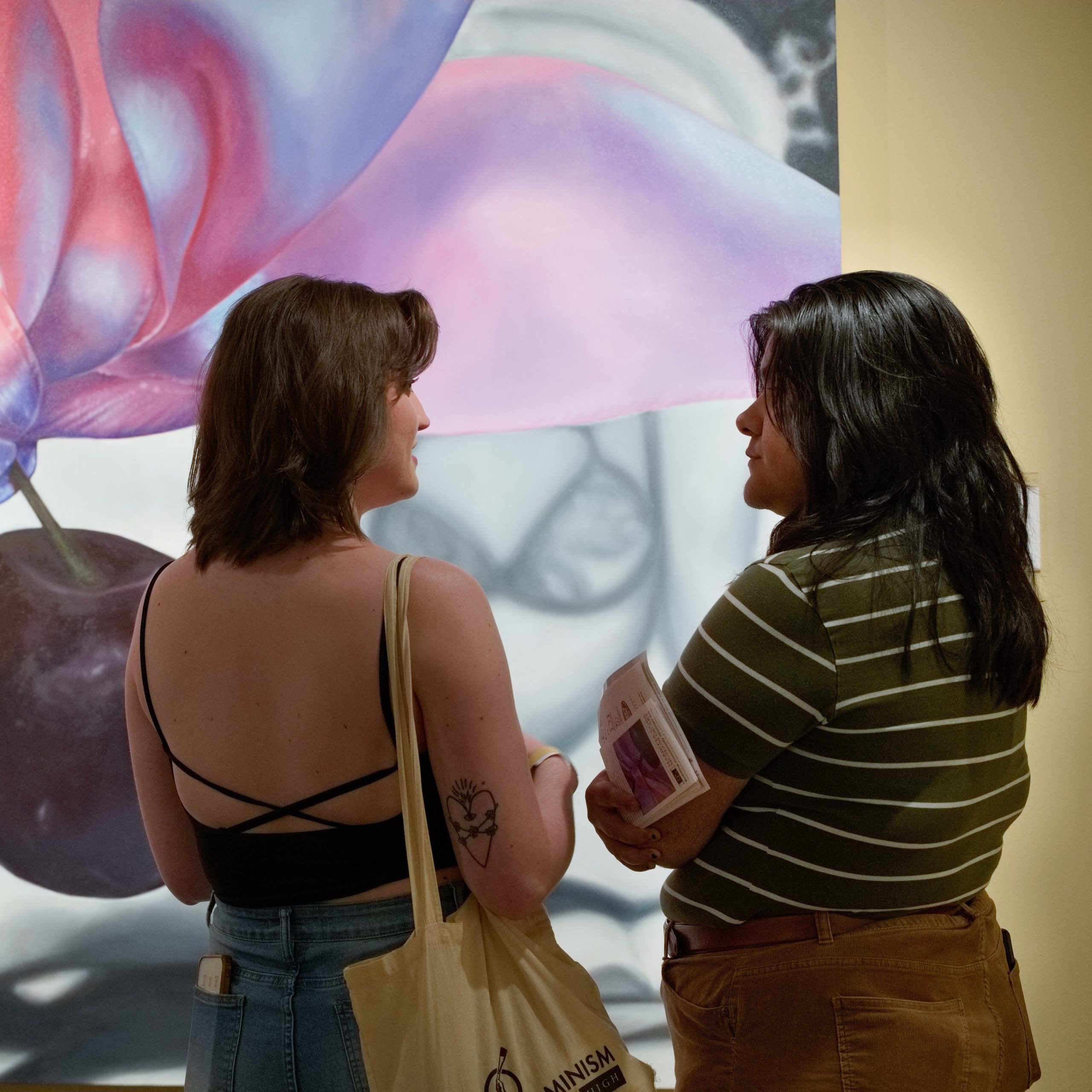 Two people seen from behind, standing side by side in front of a colorful mural; one wears a black backless top and jeans, the other a striped shirt, holding a bag and papers.
