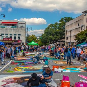 Crowd watching artists paint large colorful murals on a closed city street during a street festival on a sunny day.
