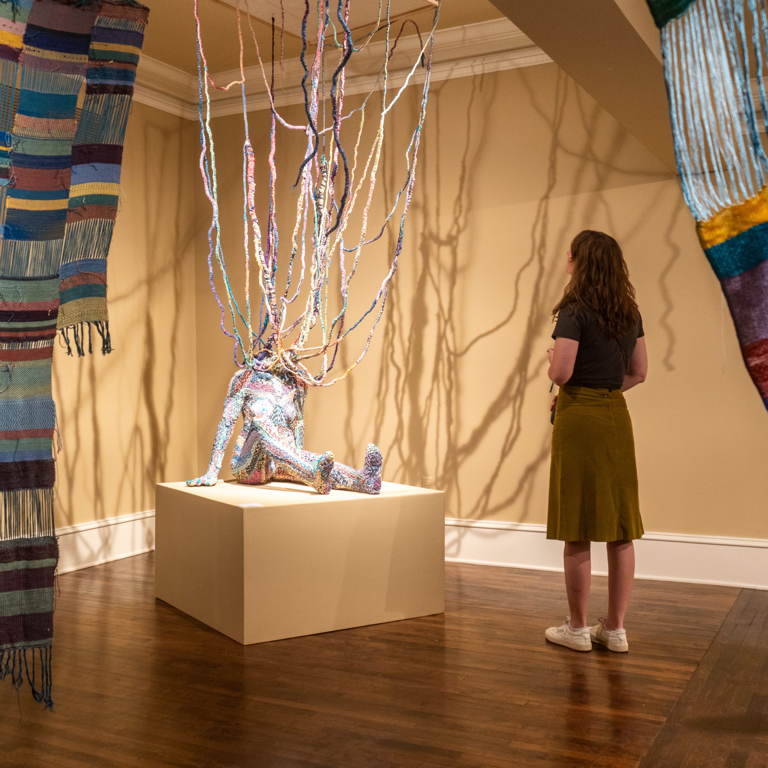 Colorful knitted strands hang from the ceiling in an art installation, with a person standing to the right.