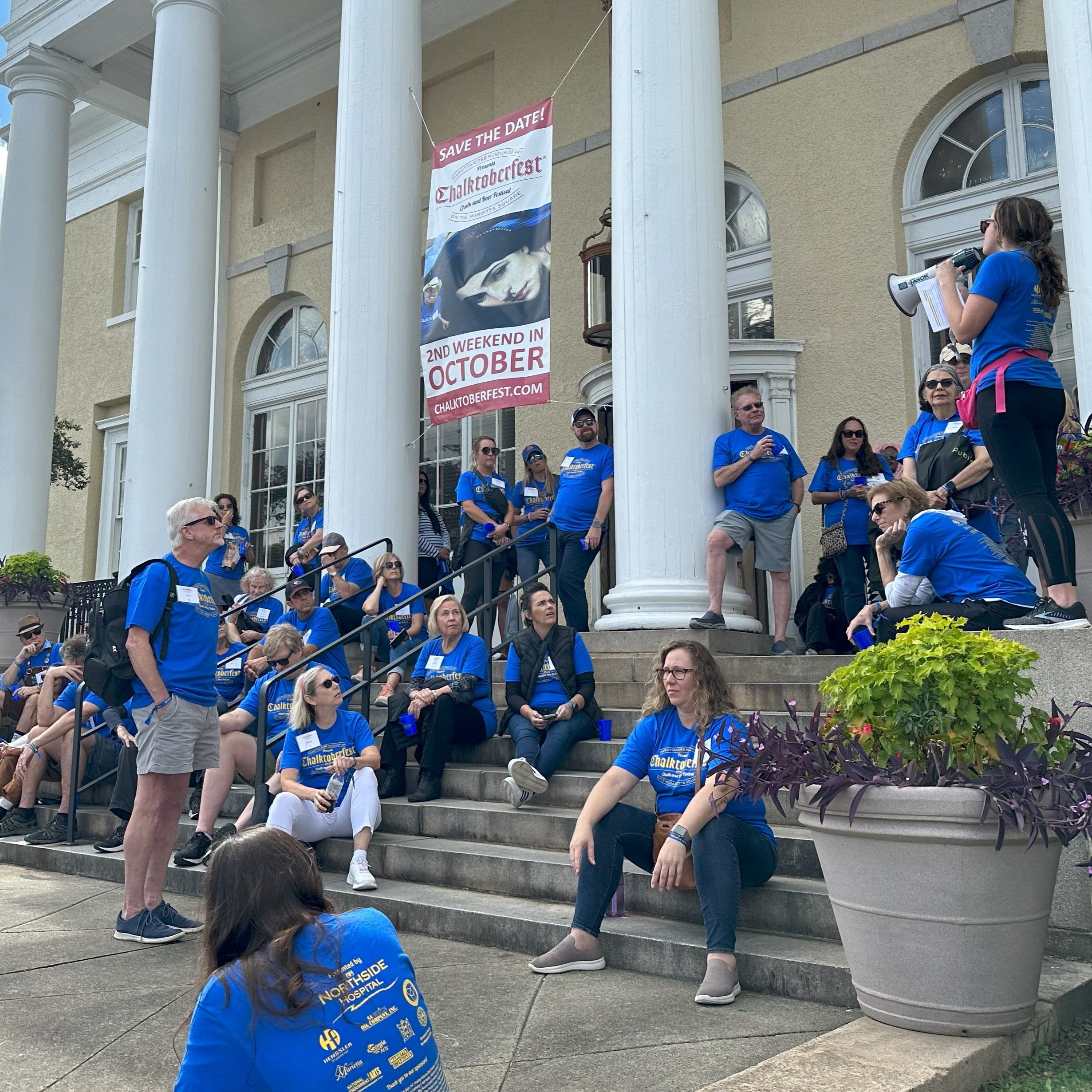 Group of people in matching blue shirts sitting on outdoor steps for an event outside a classical building with columns and banners that say Chalktoberfest.