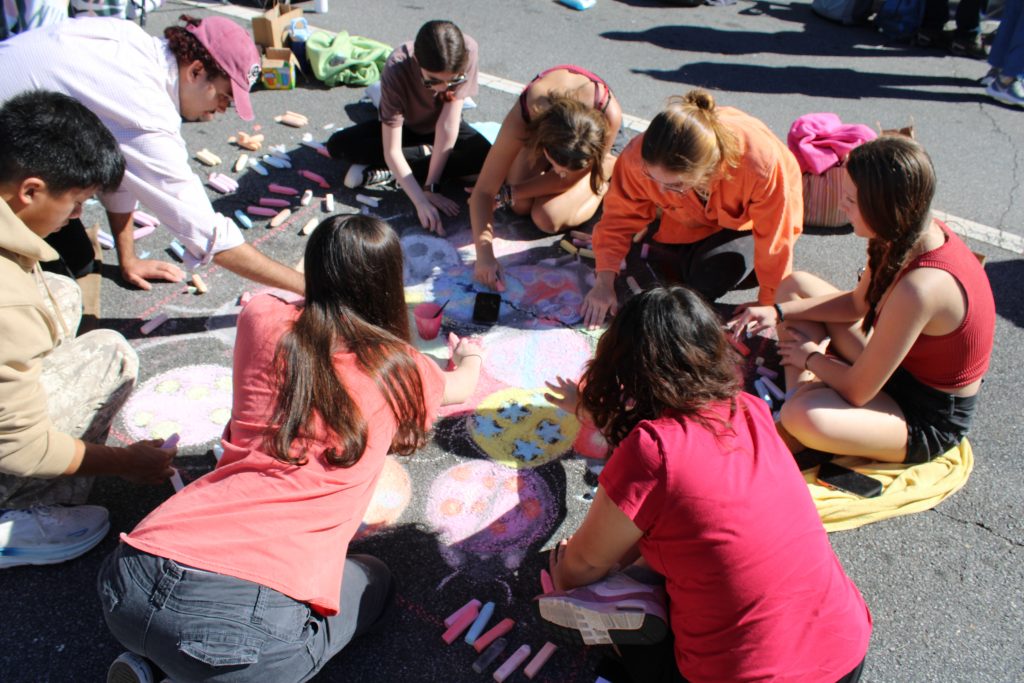 Group of people kneeling on the pavement, drawing with sidewalk chalk and colorful pieces scattered around their artwork.