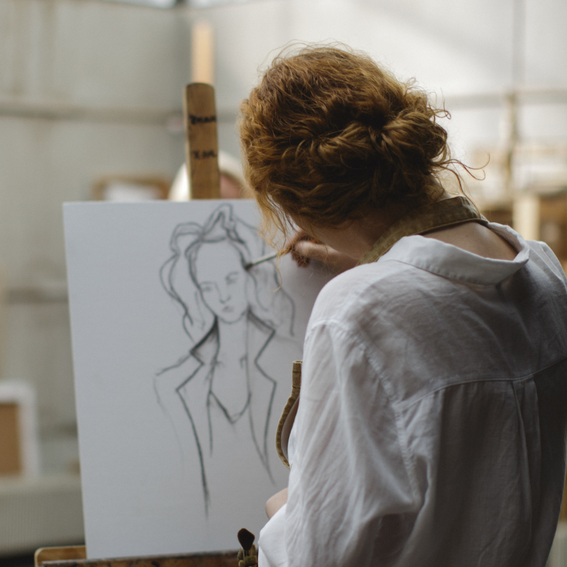 Back view of an artist with curly red hair drawing a fashion figure on a white canvas in a studio, wearing a light shirt.
