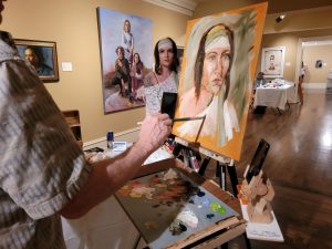Artist at an easel painting a large portrait while palette and brushes sit on a table; gallery with 2+ finished works in the background.