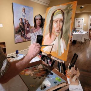 Artist at an easel painting a large portrait while palette and brushes sit on a table; gallery with 2+ finished works in the background.