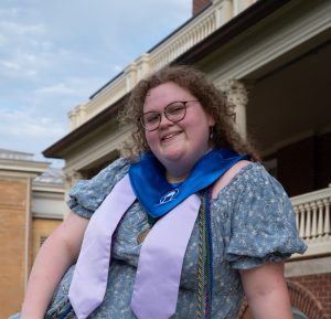 Smiling graduate in a blue gown with lavender stole and cords, standing outdoors in front of a campus building.